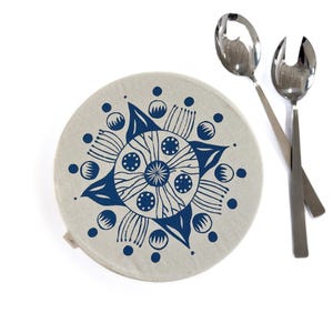 May include: A round, beige fabric bowl cover featuring a dark blue mandala-style floral print with symmetrical petals and dots, accompanied by a pair of polished stainless steel salad serving utensils with a spoon and a slotted fork design.