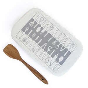 May include: A wooden spatula beside a rectangular, off-white kitchen towel. The towel has a grey pattern of kitchen utensils, including forks, knives, and spoons. The spatula is made of wood and has a smooth, curved design.