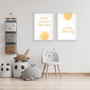 May include: Two framed prints with yellow sun illustrations and text. The left print says "here comes the sun" and the right print says "little darling". A white cat-shaped chair with a gray seat is in the foreground.