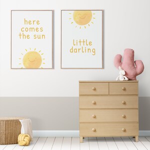 May include: Two framed prints with yellow sun illustrations and text. The left print says "here comes the sun" and the right print says "little darling". A wooden dresser with six drawers is in the foreground.