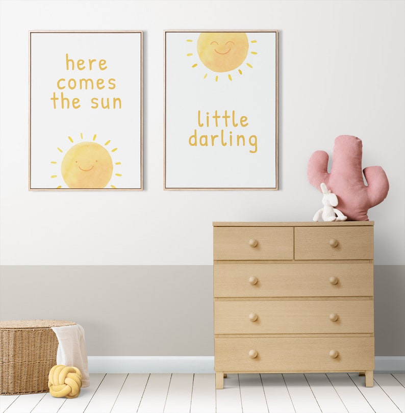 May include: Two framed prints with yellow sun illustrations and text. The left print says "here comes the sun" and the right print says "little darling". A wooden dresser with six drawers is in the foreground.