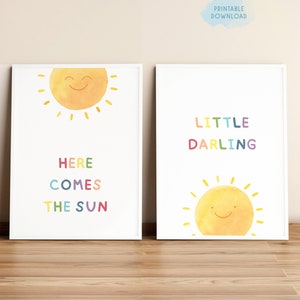 以下が含まれることがあります： 虹色のテキストで笑顔の太陽が描かれた、印刷可能な保育園の芸術作品2点。最初の作品は「Here Comes The Sun」、2番目の作品は「Little Darling」と書かれています。