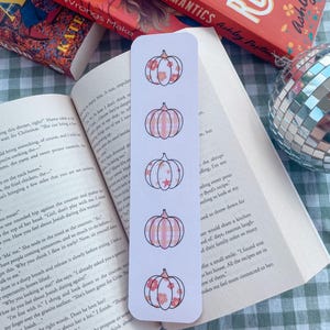 May include: A white bookmark featuring a pink and white pumpkin design. The bookmark showcases four different pumpkins with a floral pattern. This bookmark is a great way to keep your place in a book.