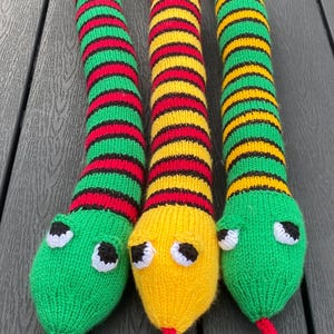 Könnte beinhalten: Drei gestrickte Schlangenspielzeuge in leuchtenden Farben. Eine Schlange ist grün, rot und schwarz, eine andere ist gelb, rot und schwarz, und die dritte ist grün, gelb und schwarz. Jede hat einen grünen oder gelben Kopf mit weißen Augen und einer roten Zunge.