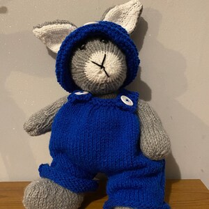 May include: A handmade knitted bunny doll wearing blue overalls and a matching hood. The bunny has gray and white fur, black eyes, and a stitched nose and mouth. The overalls have white buttons.
