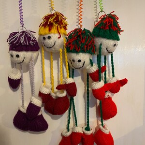 May include: Four handmade knitted ornaments hanging from a white wall. Each ornament is a white ball with a knitted hat and a pair of knitted booties. The hats are purple, yellow, green, and red. The booties are all red.
