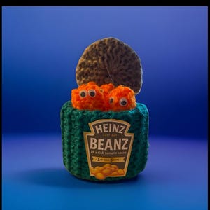 May include: A knitted Heinz Beanz can, with a green woolly cover and a brown lid. The can displays the Heinz Beanz label and contains two orange, googly-eyed bean characters. The background is a solid blue.