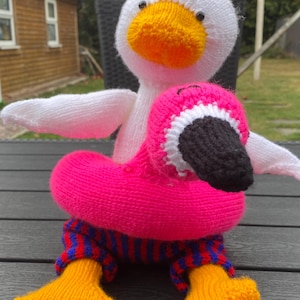 May include: A handmade knitted goose and flamingo toy. The goose is white with a yellow beak and orange feet. The flamingo is pink with a black beak and is wearing blue and red striped pants. The toys are sitting on a dark surface.