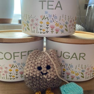 May include: White ceramic storage jars with wooden lids, labelled "TEA", "COFFEE", and "SUGAR", decorated with floral designs. A crocheted tea bag character with a smiling face and a blue tea bag.