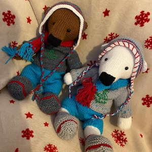 May include: Two knitted bear dolls, one brown and one white, wearing winter outfits. Each bear has a gray sweater with a green tree design, blue pants, and a matching hat and scarf. The dolls are on a cream-colored surface with red snowflake and star patterns.
