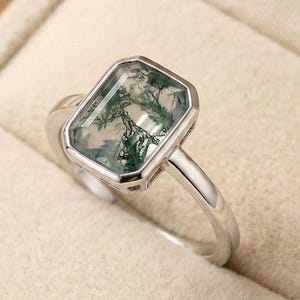 May include: A silver ring featuring a rectangular moss agate gemstone with visible green dendritic inclusions. The stone is set in a smooth metal bezel, and the band is a polished silver metal. The ring is displayed in a neutral-coloured box.