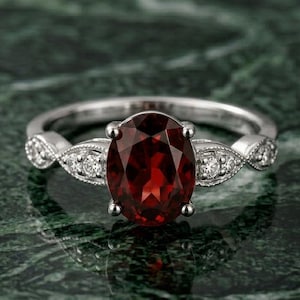 Puede incluir: Un anillo de plata con una piedra preciosa granate de color rojo intenso y corte ovalado sujeta por cuatro garras. La banda está adornada a cada lado con tres pequeños diamantes blancos redondos engastados en un marco metálico decorativo en forma de marquesa.
