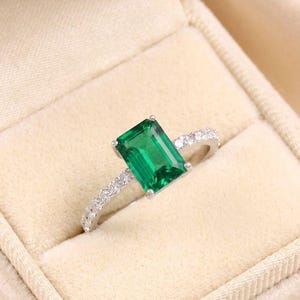 May include: A silver ring with a large, rectangular emerald-cut green gemstone as the focal point. The band is adorned with smaller, clear gemstones. The ring is set in a beige jewelry box.