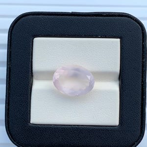 May include: A pale pink oval gemstone displayed in a black and white box. The gemstone has a faceted surface, reflecting light. The box has a black leather-like exterior and a white interior, creating a contrast that highlights the gem.