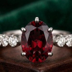 Könnte beinhalten: Ein Silberring mit einem großen, oval geschliffenen, tiefroten Granat-Edelstein, der von vier Krappen gehalten wird. Die Ringschiene ist auf jeder Seite des Mittelsteins mit drei kleinen, runden, klaren weißen Diamanten in Silber besetzt.