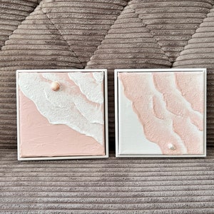 May include: Two square framed wall art pieces. Each features a textured, abstract beach scene with white waves and a pink shore. One has a seashell. The frames are white, and the background is a gray, corduroy-like fabric.