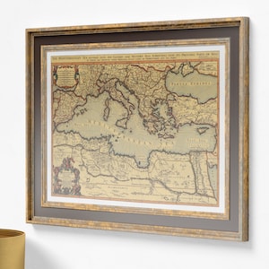 Op de afbeelding: Ingelijste vintage kaart van de Middellandse Zee, met gedetailleerde cartografie en Engelse tekst. De kaart is ingelijst in een goudkleurige houten lijst met een donkergrijze passe-partout, wat een klassieke esthetiek creëert. De kleuren van de kaart zijn gedempt, met een sepia tint.