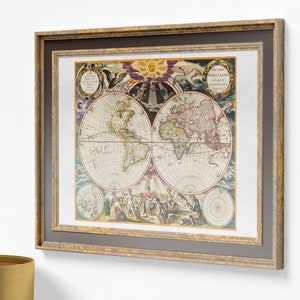 Op de afbeelding: Ingelijste vintage wereldkaart print. De kaart toont gedetailleerde illustraties van continenten en oceanen, met een focus op Amerika en Europa. De lijst is goud en grijs, wat een klassieke touch aan het kunstwerk toevoegt.
