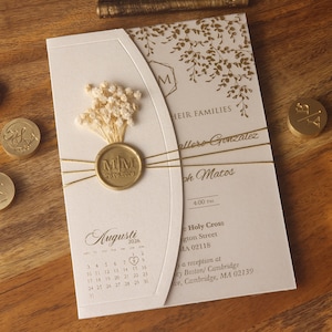 Op de afbeelding: Een crèmekleurige trouwuitnodigingskaart met een gouden waszegel met de initialen MM en de datum 08.29.2026, versierd met gedroogde gipskruidbloemen, gouden draad en een gedrukte augustus 2026 kalender met een hartje op de 29e.