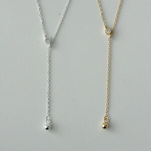 May include: Two delicate necklaces with a single small clear stone pendant and a small ball at the end of the chain. One necklace is silver and the other is gold.
