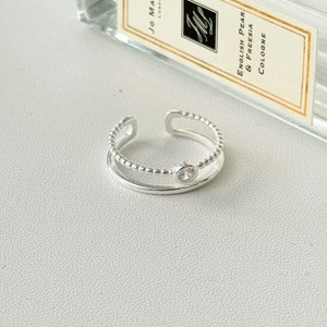 Può includere: Un anello regolabile in argento con una piccola gemma trasparente. L'anello ha una texture a perline sulla fascia esterna e una fascia interna liscia. Una bottiglia di colonia è sullo sfondo.