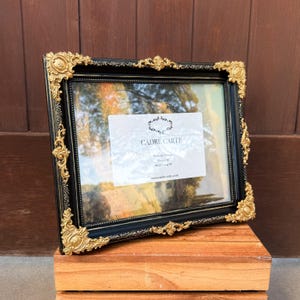May include: Black picture frame with ornate gold corner accents. The frame has a square shape and a glass front. The text "CADRE CARTE" is visible on a white label inside the frame. The frame is 8 x 10 inches.