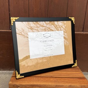 Puede incluir: Marco de fotos negro con detalles dorados en las esquinas. El marco contiene un inserto de papel con el texto "CADRE CARTE Picture Frame 10 x 8 in 25.4 x 20.32 cm". El marco está sobre una superficie de madera.