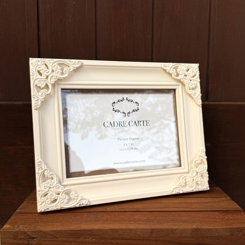 Pu&ograve; includere: Cornice per foto color crema con dettagli decorativi agli angoli. La cornice &egrave; rettangolare ed &egrave; progettata per contenere una foto. Il testo "CADRE CARTE" &egrave; visibile, insieme alle dimensioni 12,7 x 17,78 cm.