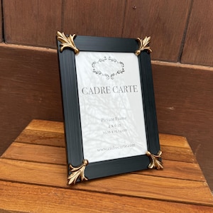 Puede incluir: Marco de fotos negro con detalles dorados en las esquinas. El marco rectangular es para una foto de 10,16 x 15,24 cm. El texto "CADRE CARTE" está impreso en el fondo blanco.