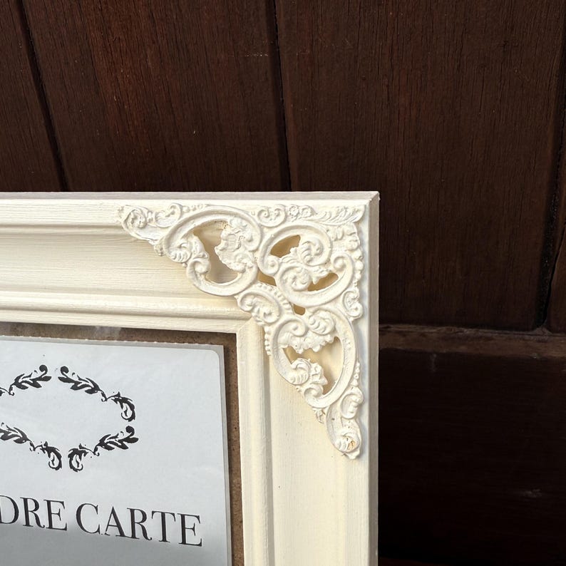 Pu&ograve; includere: Cornice color avorio con dettagli angolari ornati. La cornice ha una forma rettangolare e presenta un motivo floreale decorativo negli angoli. Le parole "DRE CARTE" sono visibili sull'immagine all'interno della cornice.