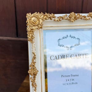 May include: A white picture frame with gold ornate detailing. The frame is 5 x 7 inches or 12.7 x 17.78 centimeters. The text "CADRE CARTE" is visible on the frame.