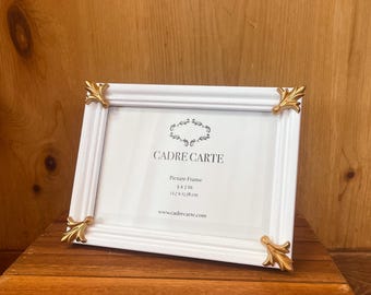 Op de afbeelding: Witte fotolijst met gouden accenten en sierlijke details. De lijst is 12,7 x 17,8 cm (5 x 7 inch) groot en heeft de tekst "CADRE CARTE" op de voorkant. Fotolijst 5 x 7 inch 12,7 x 17,8 cm www.cadrecarte.com