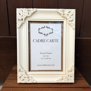 Pu&ograve; includere: Cornice color crema con dettagli angolari ornati. La cornice presenta il testo "CADRE CARTE" e le dimensioni "12,7 x 17,78 cm". La cornice &egrave; realizzata in materiale solido ed &egrave; esposta su una superficie di legno.