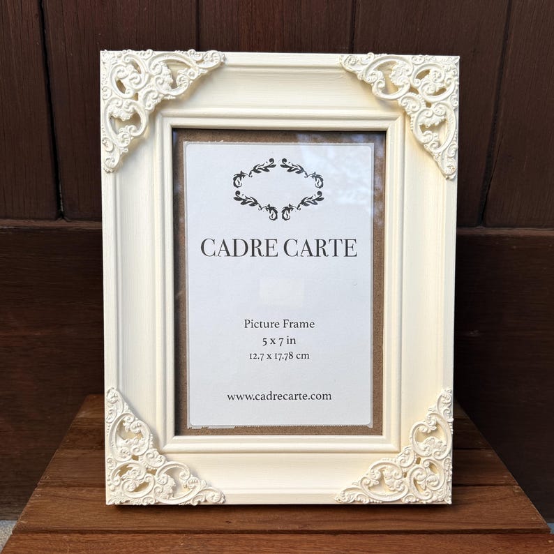 Pu&ograve; includere: Cornice color crema con dettagli angolari ornati. La cornice presenta il testo "CADRE CARTE" e le dimensioni "12,7 x 17,78 cm". La cornice &egrave; realizzata in materiale solido ed &egrave; esposta su una superficie di legno.