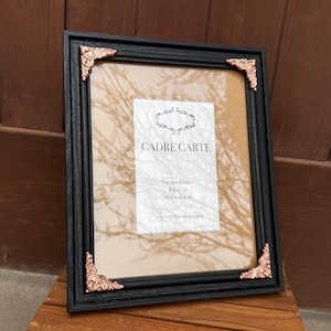 May include: A black rectangular picture frame with ornate copper-colored metal corner accents. The insert displays the text CADRE CARTE, Picture Frame, 8 x 10 in, 20.32 x 25.4 cm, and the website address www.cadrecarte.com.