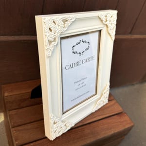 Pu&ograve; includere: Cornice per foto color avorio con dettagli decorativi agli angoli. Contiene una carta bianca con il testo "CADRE CARTE" e "Picture Frame 5x7 in 12.7 x 17.8 cm". La cornice &egrave; su una superficie di legno.