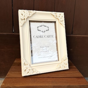 Pu&ograve; includere: Cornice color avorio con dettagli decorativi agli angoli. La cornice &egrave; etichettata "CADRE CARTE" e misura 12,7 x 17,78 cm. La cornice &egrave; esposta su una superficie di legno.