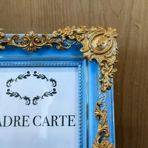 Puede incluir: Un marco de fotos azul con detalles dorados. El marco tiene forma rectangular con esquinas doradas ornamentadas y el texto "ADRE CARTE". El marco es de madera y tiene un acabado envejecido.