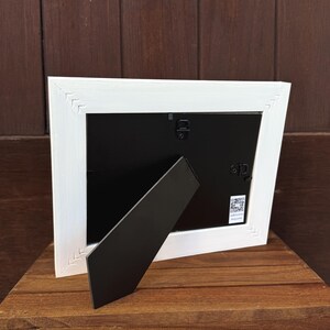 Pu&ograve; includere: Una cornice bianca con retro nero e cavalletto nero. La cornice ha un design strutturato a forma di chevron sugli angoli. La cornice &egrave; su una superficie di legno.
