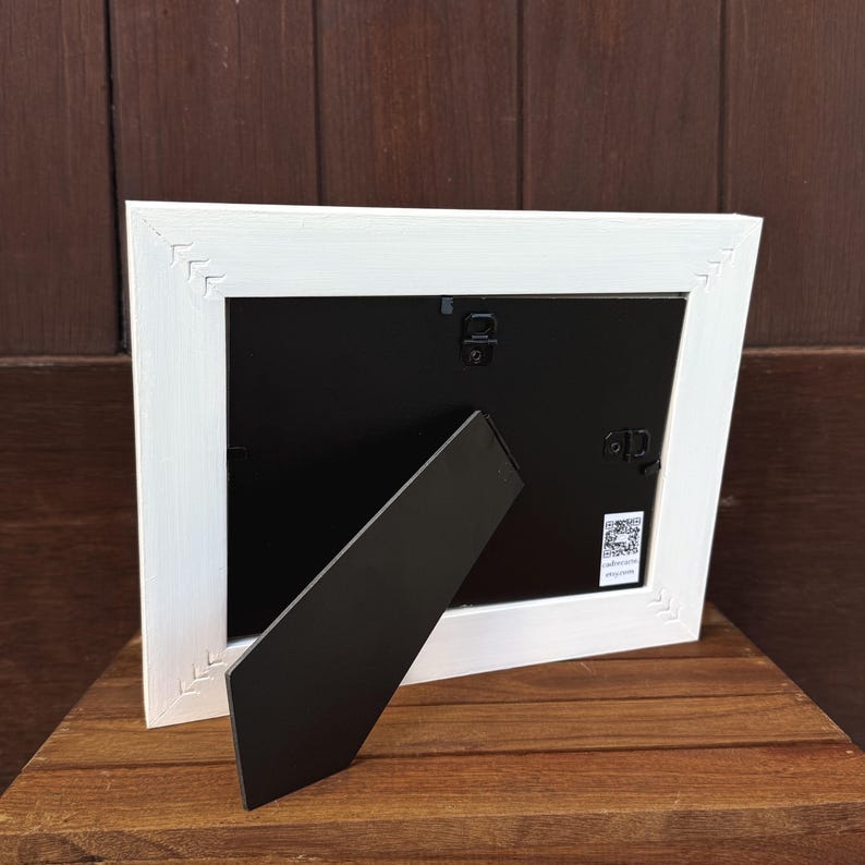Pu&ograve; includere: Una cornice bianca con retro nero e cavalletto nero. La cornice ha un design strutturato a forma di chevron sugli angoli. La cornice &egrave; su una superficie di legno.