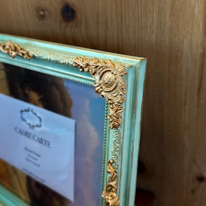 May include: A close-up of a rectangular picture frame with a light teal finish and ornate gold corner accents. The frame holds a picture with a blue sky background. The text "CADRECARTE" is visible on the picture.