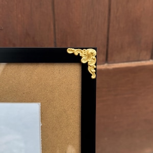 Könnte beinhalten: Schwarzer Bilderrahmen mit einer goldenen Zierecke. Der Rahmen hat einen schwarzen Rand und eine hellbraune Rückseite. Die goldene Ecke ist verziert und verleiht dem Rahmen einen Hauch von Eleganz.
