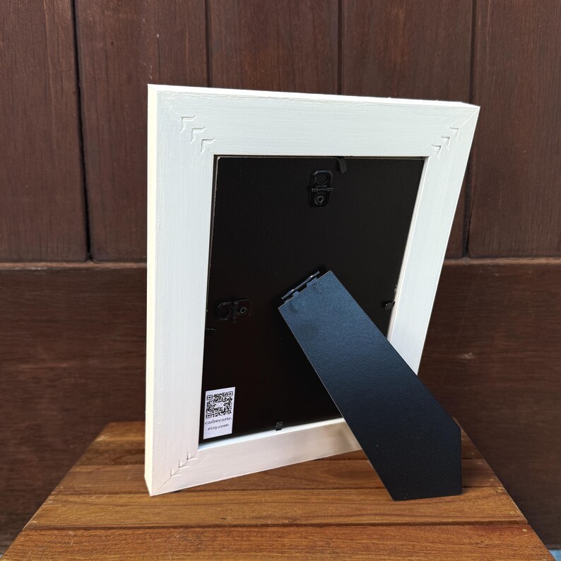 Pu&ograve; includere: Una cornice bianca con retro nero e cavalletto nero. La cornice ha un design decorativo negli angoli. La cornice &egrave; appoggiata su una superficie di legno.