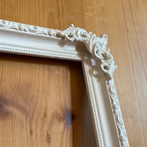 Puede incluir: Marco de fotos marfil adornado con detalles de volutas y acentos decorativos en las esquinas. El marco tiene una estética clásica y vintage, perfecto para exhibir fotografías o obras de arte. El marco está hecho de un material sólido.