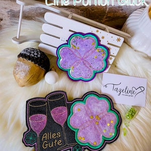 May include: Two purple and green embroidered patches with a four-leaf clover design. One patch says "Eine Portion Glück" and the other says "Alles Gute" with two wine glasses.