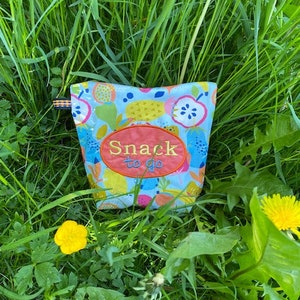 Könnte beinhalten: Eine blaue und weiße Stoff-Snacktasche mit einem bunten Früchte-Druck und dem Text "Snack to go", der in Orange auf einem roten Oval gestickt ist.