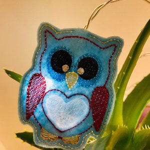 May include: A blue felt owl with a red heart and gold stitching. The owl is hanging from a string of lights.