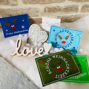 Könnte beinhalten: Weihnachtsdekorationen aus Filz in Blau, Grün und Hellblau mit Rentier-Designs und Text. Ein weißes Holzschild mit der Aufschrift "love" und ein herzförmiger Schmuck sind ebenfalls zu sehen.