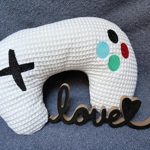 May include: White, waffle-textured pillow shaped like a video game controller, with a black cross and coloured buttons. A wooden cutout spelling "love" is placed in front of the pillow. The pillow is on a grey surface.