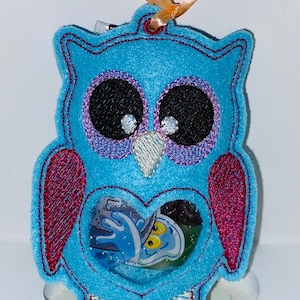 May include: A blue felt owl ornament with a heart-shaped window revealing a blue and yellow ghost inside. The owl has pink wings and embroidered eyes.